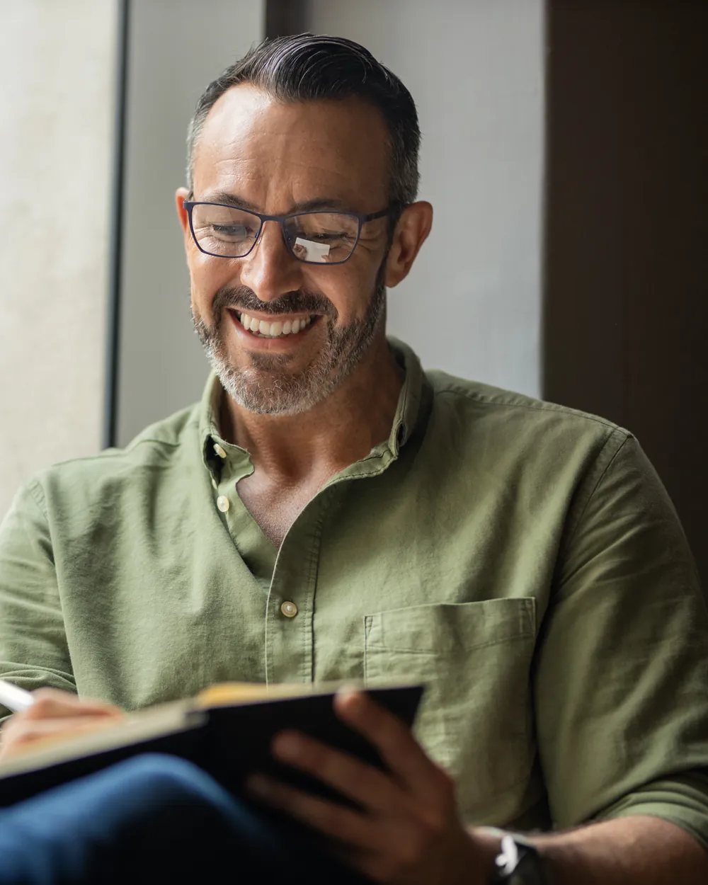 A man smiles to himself as he writes in a notebook by a window