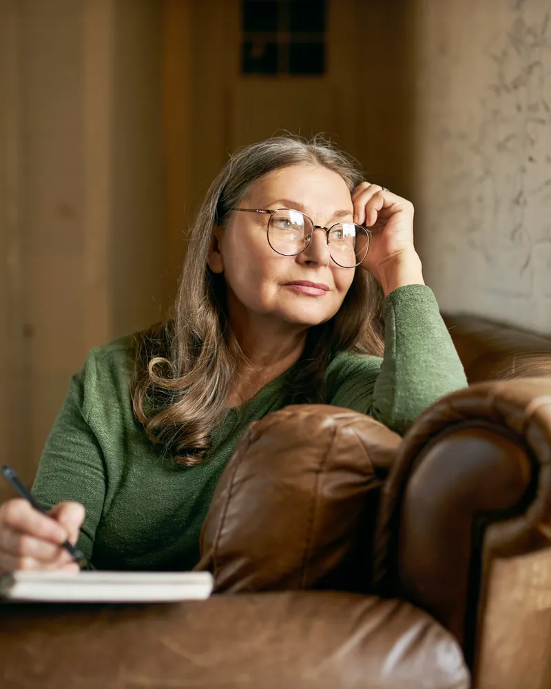 A woman in glasses pauses mid-thought while writing in her favorite chair