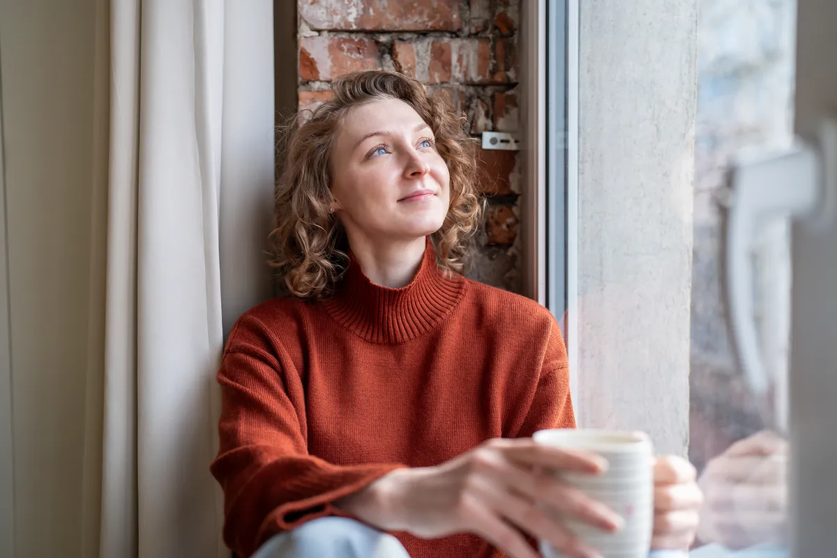 A woman sits by a window with a cup of coffee, looking out